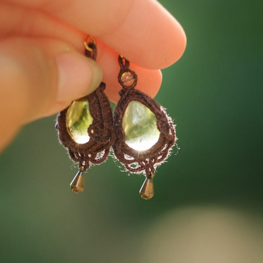 Boucles d'oreilles de style celtique elfique. Bijou féminin composé de : un tissage en relief avec arabesques en micro macramé avec du fil de coton marron foncé chocolat, pierre naturelle de citrine de forme goutte couleur jaune miel accompagné d'une perle ronde en citrine de 3 mm, d'un crochet doré en acier et d'une micro goutte bronze. Le bijou est présenté tenu dans une main avec une transparence évidente et un rayon de soleil qui traverse la pierre
