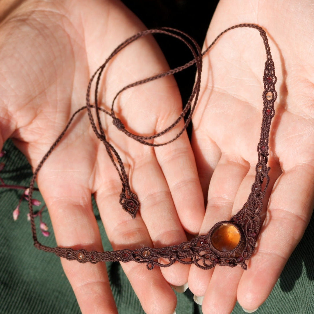 Collier ras-de-cou elfiques en micro macramé avec pierre naturelle. Bijoux tissé en fil de coton de petit diamètre. Mini Grenats rouge rosé facettés autour d'une citrine jaune ambré, fil de coton marron foncé. Motifs tissés en entrelacs celtiques et arabesques elfiques.système à coulisse tubulaire et embout tissé. Bijoux ambiance bohème hippie aux couleurs chaudes. Bijoux posés dans 2 mains en coupe, vue d'en haut dans une main à la peau claire