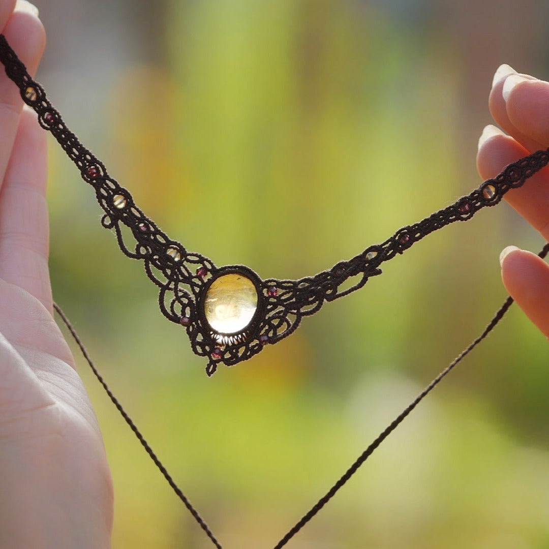 Collier ras-de-cou elfiques en micro macramé avec pierre naturelle. Bijoux tissé en fil de coton de petit diamètre. Mini Grenats rouge rosé facettés autour d'une citrine jaune ambré ovale, fil de coton marron foncé. Motifs tissés en entrelacs celtiques et arabesques elfiques Forme V. Bijoux ambiance bohème hippie aux couleurs chaudes. Bijoux tenu à la verticale avec fond claire et nature. Bijoux vue de face avec transparence et jeu de lumière