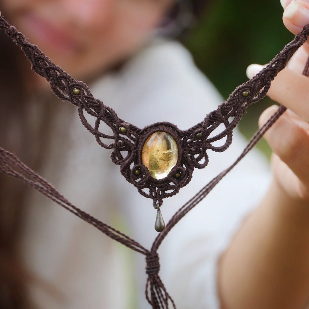Collier large en micro macramé de couleur marron foncé, jaune ambré et bronze. Motif tissé en micro macramé avec ambiance entrelacs celtiques et arabesques elfiques. Présence d'une pierre centrale ovale de grande taille jaune translucide, gouttes bronze, forme V avec mini perles ronde bronze parsemées. Photo ambiance bohème hippie avec une touche d'élégance. Bijou tenu en verticale avec transparence de la pierre évidente, chemise blanche en arrière plan flou