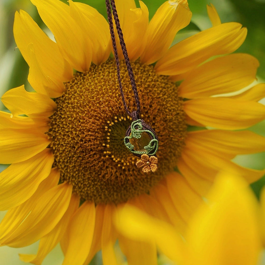 Collier avec pendentif en micro macramé avec motifs floraux. Bijou féminin composé d'un citrine jaune pâle translucide ovale sertie dans une coque marron foncé tissé avec un fil de 0.5 mm sur lequel est fixé une liane verte entrelacée avec mini feuilles et une fleur à 5 pétales jaune orangé avec un cœur doré de 2 mm.
Le bijou est en verticale. Le bijou est pris de face posé sur une fleur de tournesol jaune vif. Fond vert naturel
