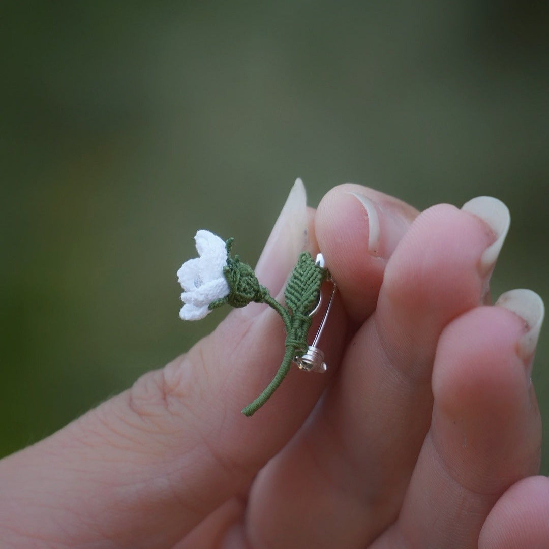 Broche miniature florale blanche en micro macramé avec labradorite bleu-vert au centre, tenue dans une main sur fond de feuilles vertes, bijou bohème et elfique artisanal.