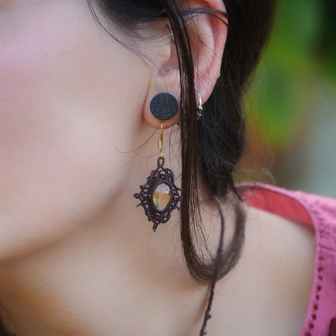 Boucles d'oreilles elfiques en micro macramé avec pierre naturelle. Bijoux tissé en fil de coton de petit diamètre. 4 Mini Grenats rouge rosé facettés autour d'une citrine jaune ambré, fil de coton marron foncé. Motifs tissés en entrelacs celtiques et arabesques elfiques. Crochet doré. Bijou ambiance bohème hippie aux couleurs chaudes. Bijou porté, vue de 3/4 sur un modèle féminin à la peau claire et aux cheveux bruns. Habits rose font vert