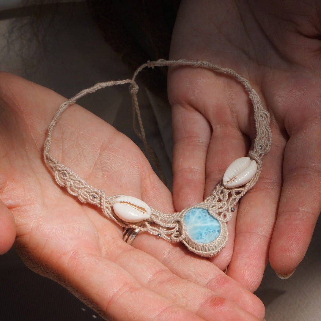 Collier plastron en Larimar Océan bleu ciel, coton couleur lin naturel (beige gris), cauris blancs et perles en acier inoxydable argenté. Système de réglable en tube tissé à l'arrière du bijou. Tissage symétrique en micro macramé avec effet nœuds celtiques entrelacés en relief. Style bohème, hippie, plage, Sirène, tribal et tropical. Convient pour un mariage type bohème ou plage. Bijou tenu vue 3/4, tenue romantique et bohème blanche avec dentelle apparente. Modèle à la peau bronzée et aux cheveux bruns.