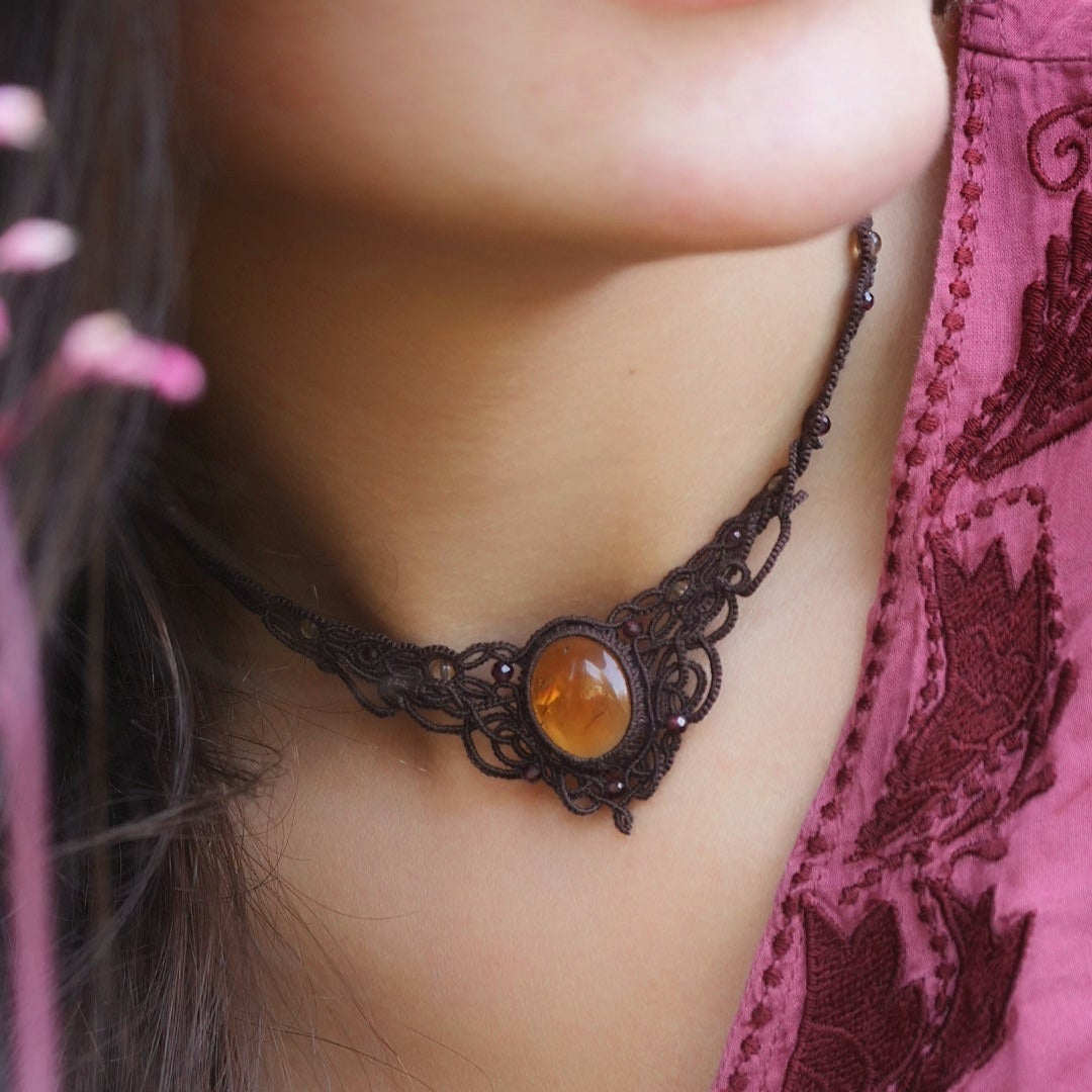 Collier ras-de-cou elfiques en micro macramé avec pierre naturelle. Bijoux tissé en fil de coton de petit diamètre. Mini Grenats rouge rosé facettés autour d'une citrine jaune ambré ovale, fil de coton marron foncé. Motifs tissés en entrelacs celtiques et arabesques elfiques Forme V. Bijoux ambiance bohème hippie aux couleurs chaudes. Bijoux porté sur un mannequin féminin à la eau claire et cheveux bruns, habit rose fuchsia. Vue de face
