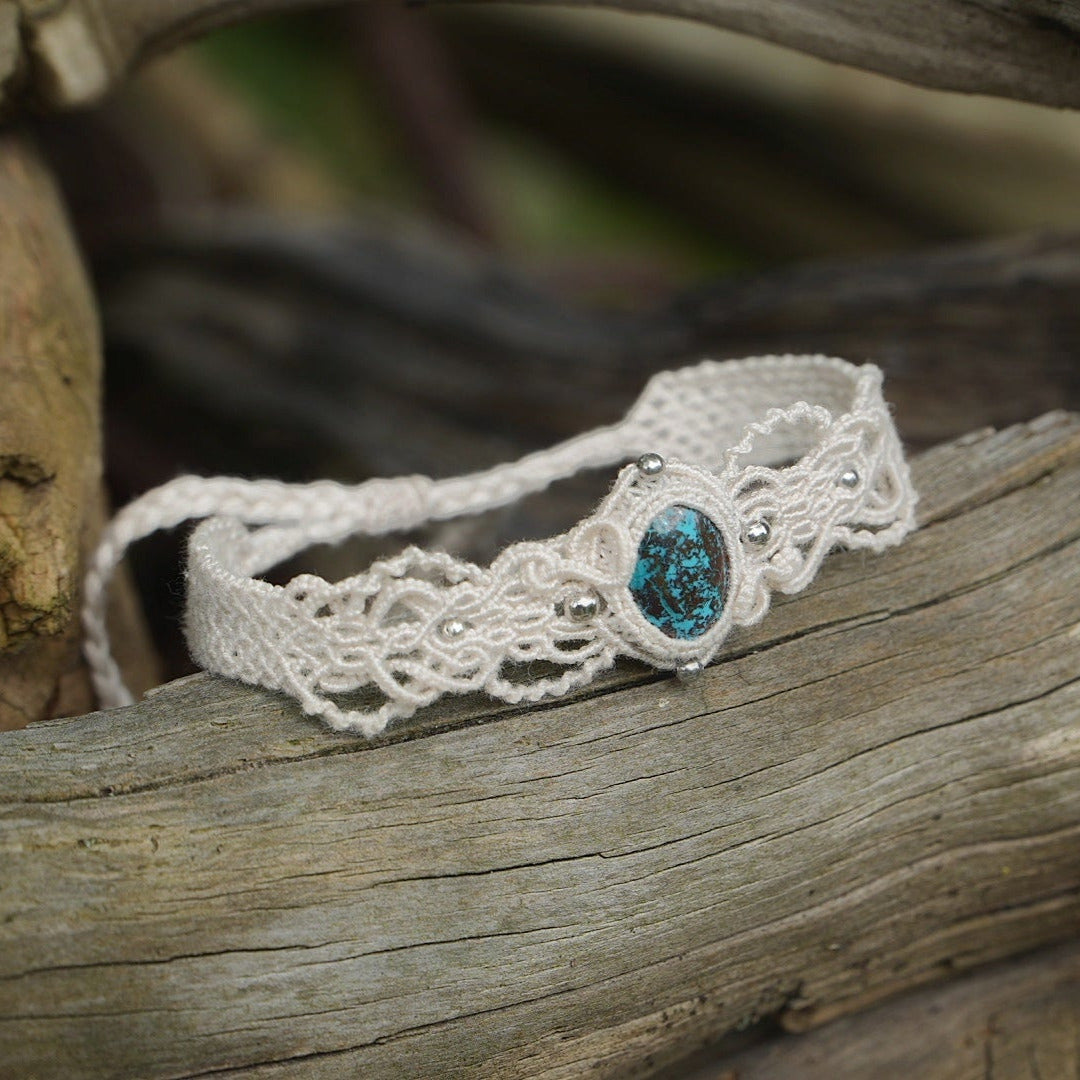 Bracelet large asymétrique. Bijou dentelle ambiance bohème hippie. Bracelet tissé en micro macramé avec fil de coton écru de 0.5 mm avec une pierre ovale de chrysocolle couleur bleu turquoise et marron foncé chocolat. Présence de part et d'autre de façon symétrique des perles en acier inoxydable argentées. Système de réglage à coulisse tubulaire à l'arrière. Design en relief et en torsade entrelacées celtiques. Ambiance plages, festival d'été et mariage boho. Bijou posé sur un galet beige et coquillages.