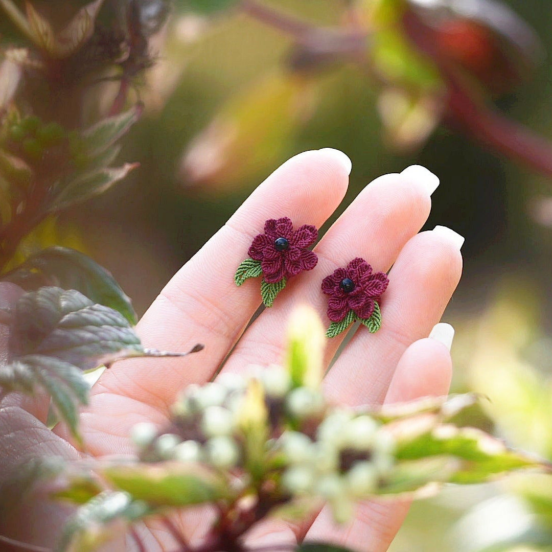 Boucles d’oreilles florales en micro macramé faites main, collection « Pétales éternels », posées sur une main féminine. Fleur 3D tissée en coton rouge bordeaux et vert tendre avec 13 pétales, 2 feuilles et cœur en perle d’onyx noire. Style bohème, elfique et hippie, idéal pour festivals et costumes féériques, photographié au milieu de feuilles et de nature verdoyante.
