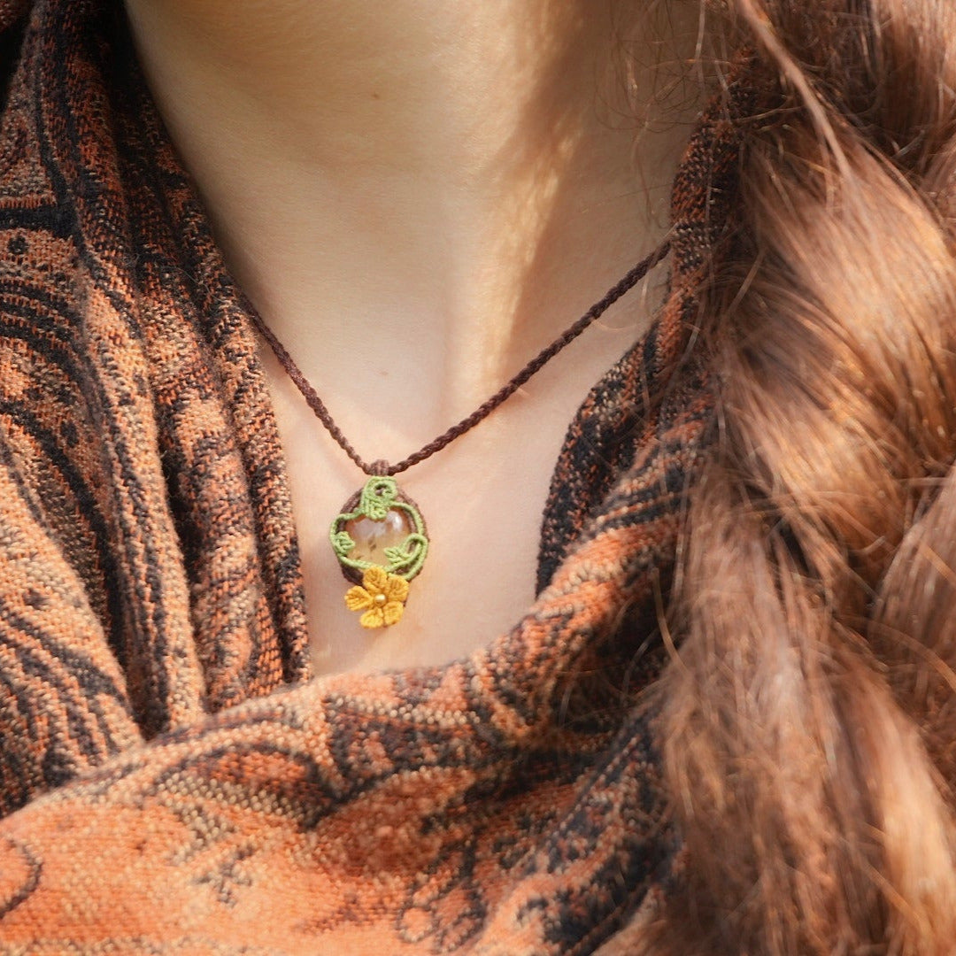 Collier avec pendentif en micro macramé avec motifs floraux. Bijou féminin composé d'un citrine jaune pâle translucide ovale sertie dans une coque marron foncé tissé avec un fil de 0.5 mm sur lequel est fixé une liane verte entrelacée avec mini feuilles et une fleur à 5 pétales jaune orangé avec un cœur doré de 2 mm.
Le bijou est porté autour d'un cou de femme à la peau claire et d'un châle orange et noir et natte brune. le bijou est pris de face avec rayon du soleil
