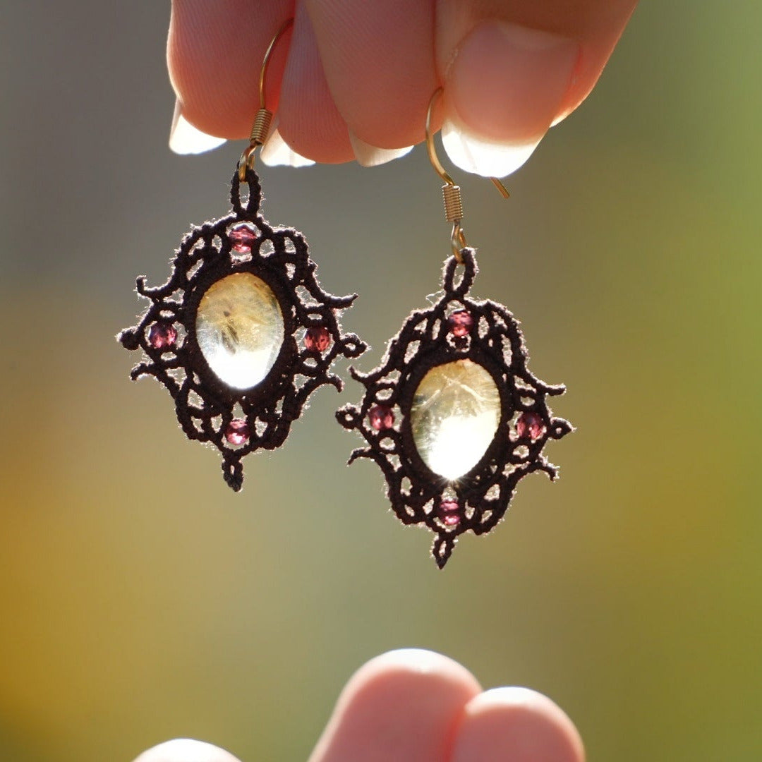 Boucles d'oreilles elfiques en micro macramé avec pierre naturelle. Bijoux tissé en fil de coton de petit diamètre. 4 Mini Grenats rouge rosé facettés translucides autour d'une citrine jaune ambré translucide, fil de coton marron foncé. Motifs tissés en entrelacs celtiques et arabesques elfiques. Crochet doré. Bijou ambiance bohème hippie aux couleurs chaudes. Bijou tenu dans une mains de façon verticale, prise de vue de face avec un jeu de lumière de façon contrejour