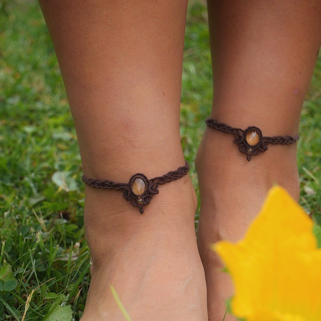 Bijoux de pieds, paire assortie de bracelets de chevilles à ambiance bohème celtique elfique. Fil marron foncé fin tissé en micro macramé avec entrelacs et arabesques, pierre centrale ovale petite taille jaune miel translucide. Bijoux hippie, bohème avec inspiration nature. Chevillères portées sur des chevilles à la peau pâle, bijoux de face avec fleurs jaune de courges au premier plan, fond herbe verte
