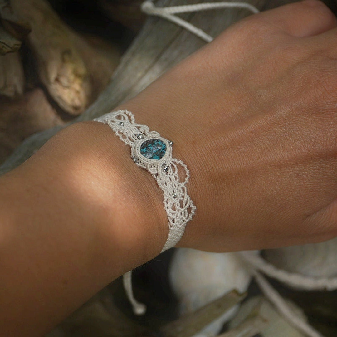 Bracelet large asymétrique. Bijou dentelle ambiance bohème hippie. Bracelet tissé en micro macramé avec fil de coton écru de 0.5 mm avec une pierre ovale de chrysocolle couleur bleu turquoise et marron foncé chocolat. Présence de part et d'autre de façon symétrique des perles en acier inoxydable argentées. Système de réglage à coulisse tubulaire à l'arrière. Design en relief et en torsade entrelacées celtiques. Ambiance plages, festival d'été, tribal. Bijou porté fond bois clair