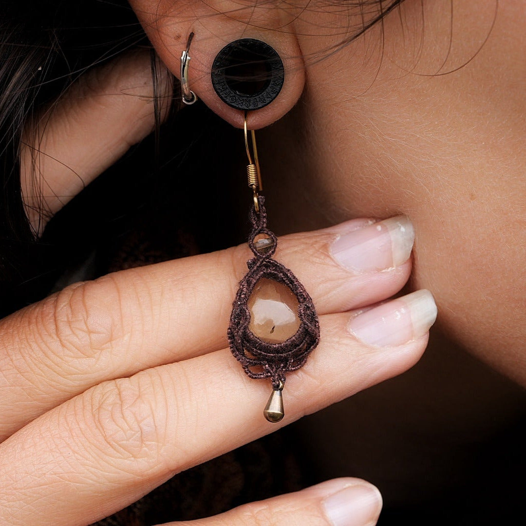 Boucles d'oreilles de style celtique elfique. Bijou féminin composé de : un tissage en relief avec arabesques en micro macramé avec du fil de coton marron foncé chocolat, pierre naturelle de citrine de forme goutte couleur jaune miel accompagné d'une perle ronde en citrine de 3 mm, d'un crochet doré en acier et d'une micro goutte bronze. Le bijou est porté sur une peau dorée et avec un fond de cheveux brun foncé.
