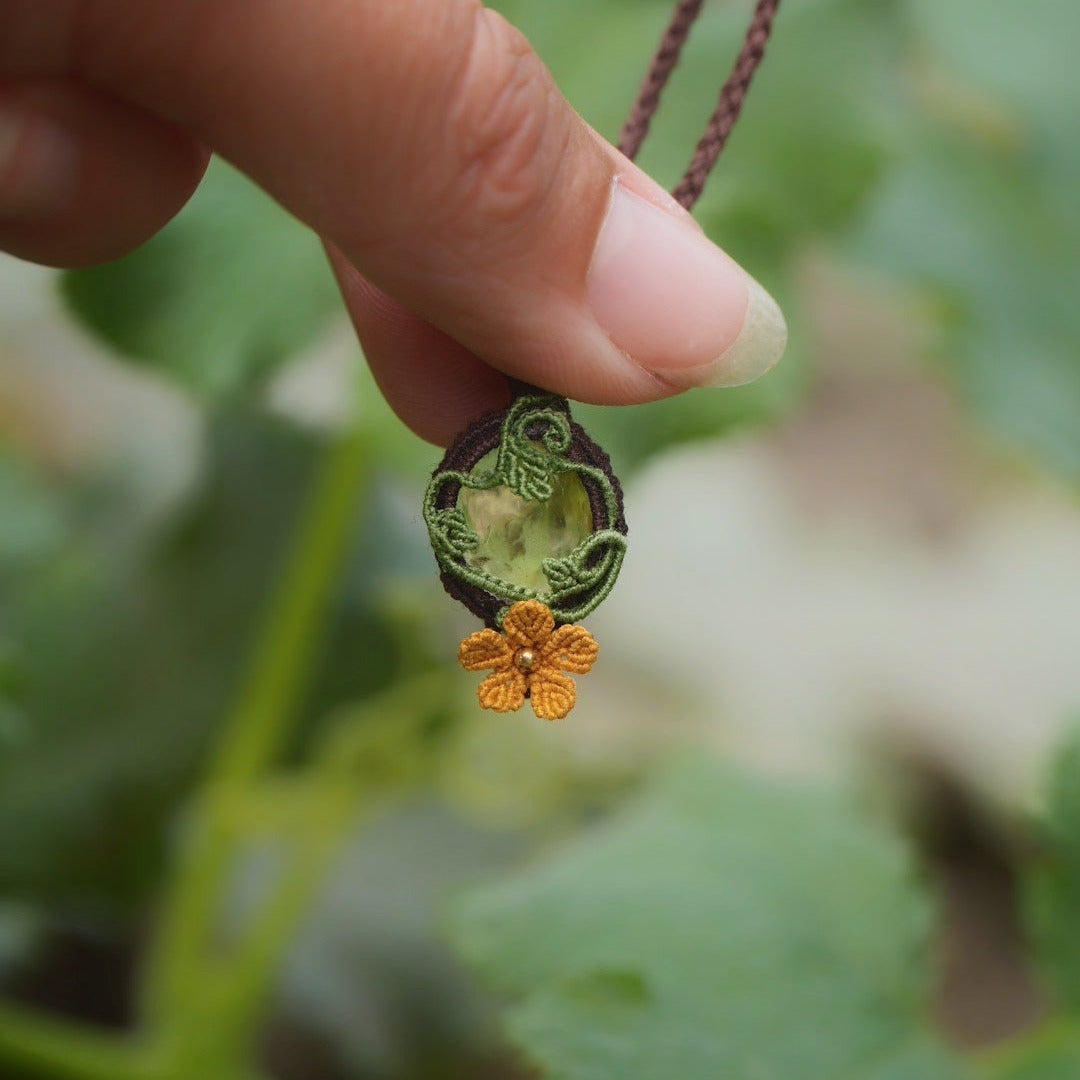 Collier avec pendentif en micro macramé avec motifs floraux. Bijou féminin composé d'un citrine jaune pâle translucide ovale sertie dans une coque marron foncé tissé avec un fil de 0.5 mm sur lequel est fixé une liane verte entrelacée avec mini feuilles et une fleur à 5 pétales jaune orangé avec un cœur doré de 2 mm.
Le bijou est en verticale. Le bijou est pris de 3/4 avec une certaine transparence et un fond vert naturel, au milieu des tortillons et feuilles de courges