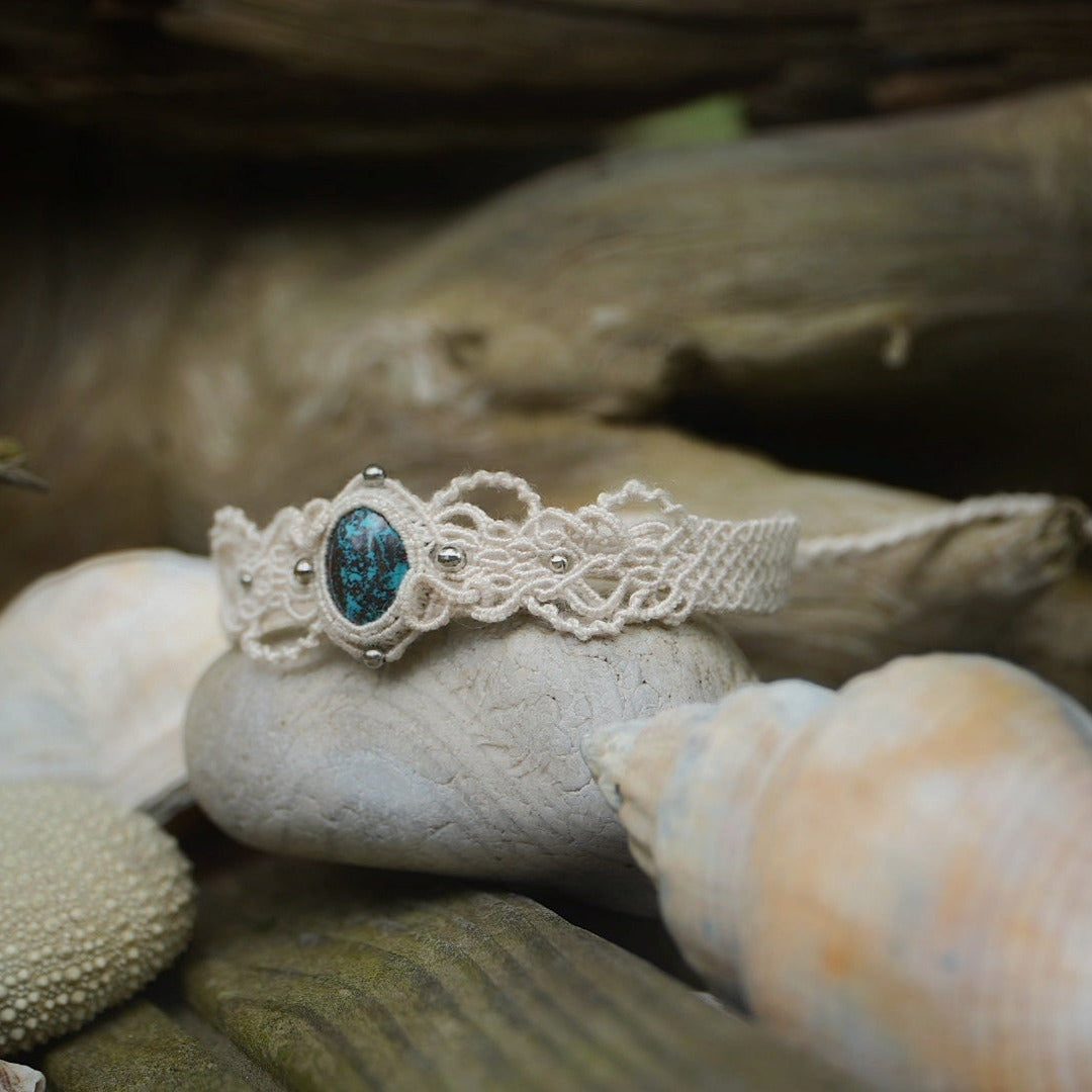 Bracelet large asymétrique. Bijou dentelle ambiance bohème hippie. Bracelet tissé en micro macramé avec fil de coton écru de 0.5 mm avec une pierre ovale de chrysocolle couleur bleu turquoise et marron foncé chocolat. Présence de part et d'autre de façon symétrique des perles en acier inoxydable argentées. Système de réglage à coulisse tubulaire à l'arrière. Design en relief et en torsade entrelacées celtiques. Ambiance plages, festival d'été et mariage boho. Bijou posé sur un galet beige et coquillages.
