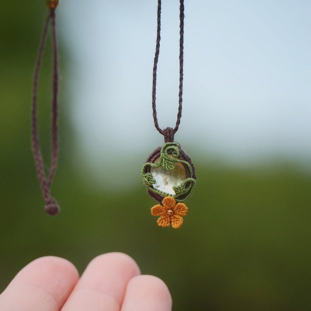 Collier avec pendentif en micro macramé avec motifs floraux. Bijou féminin composé d'un citrine jaune pâle translucide ovale sertie dans une coque marron foncé tissé avec un fil de 0.5 mm sur lequel est fixé une liane verte entrelacée avec mini feuilles et une fleur à 5 pétales jaune orangé avec un cœur doré de 2 mm.
Le bijou est en verticale, tenu dans le vide. le bijou est pris de face avec une certaine transparence et un fond vert naturel