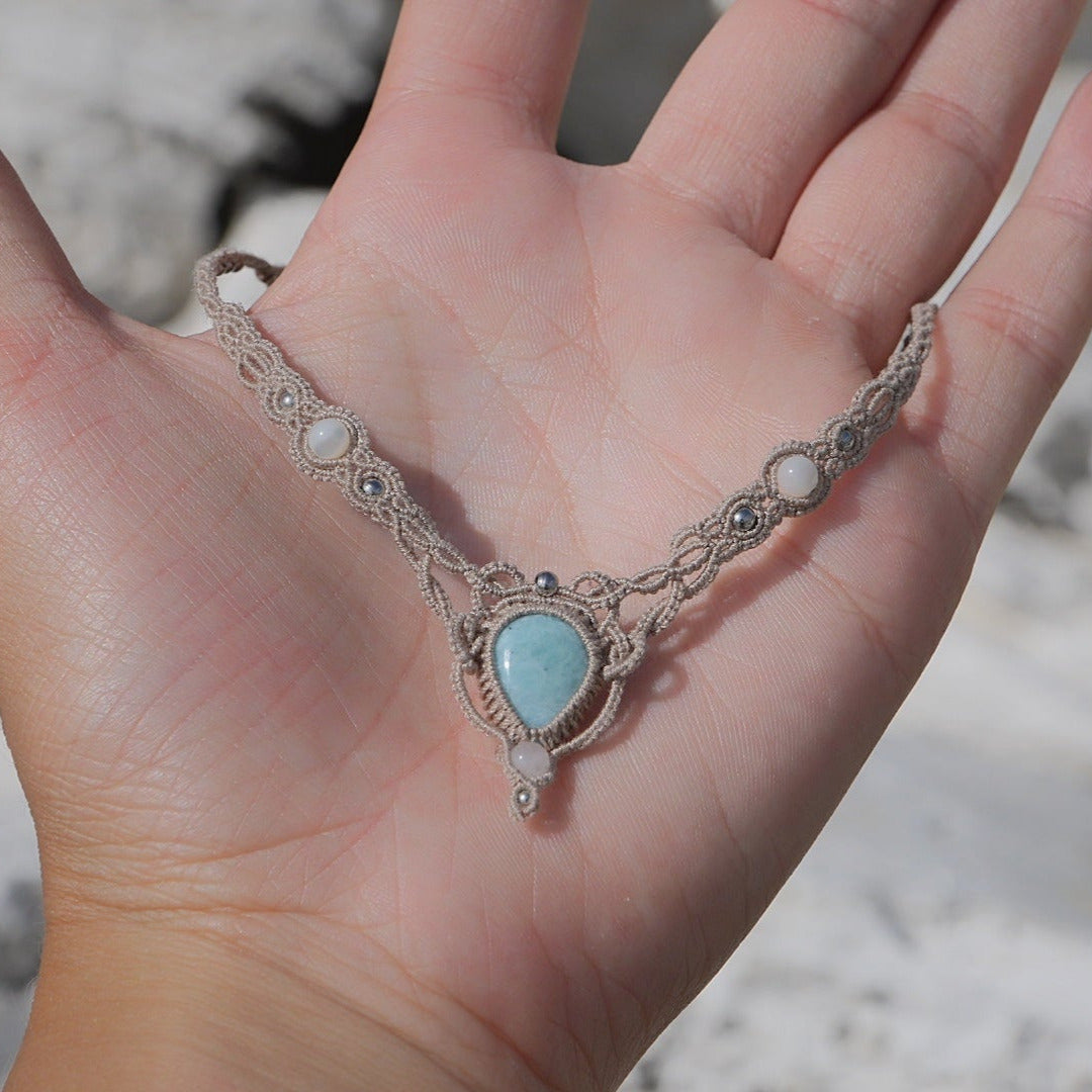 Collier ras-de-cou forme v Trésors d'Ecume composée d'une Larimar bleu ciel en poire pointe en bas, perles en nacre blanche ronde rappel des coquillages et de la plage, mini Perles rondes en acier inoxydable argenté pour le côté luxe et précieux. Fil de Coton couleur lin naturel (beige gris) de 0, 5 mm tissé en micro-macramé en relief version celtique. Collage 3 photos : Bijoux tenu à plat dans une main à la peau blanche. Photo ambiance tropiques et plages, style bohème hippie