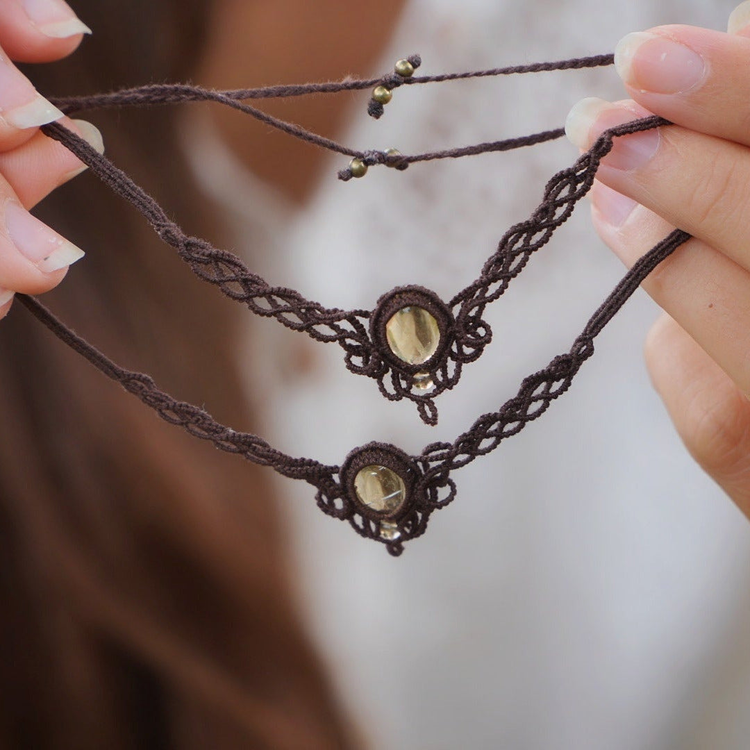 Bijoux de pieds, paire assortie de bracelets de chevilles à ambiance bohème celtique elfique. Fil marron foncé fin tissé en micro macramé avec entrelacs et arabesques, pierre centrale ovale petite taille jaune miel translucide. Bijoux hippie, bohème avec inspiration nature. Chevillères tenues entre 2 mains à la peau pâle l'une au dessus de l'autre en verticale avec en fond une femme aux cheveux bruns et avec une chemise blanche