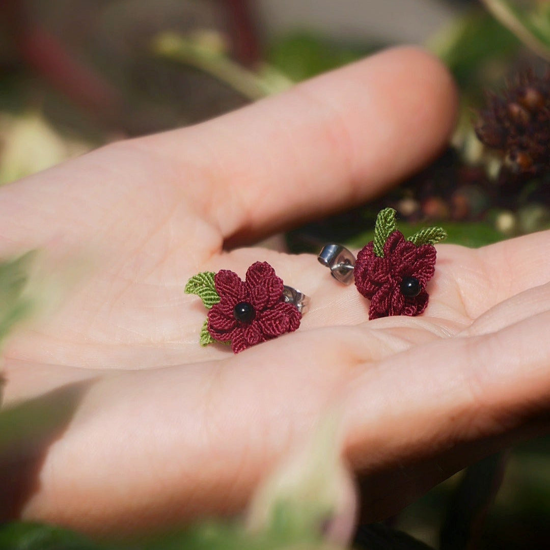 Boucles d’oreilles florales en micro macramé faites main, collection « Pétales éternels », posées sur une main féminine. Fleur 3D tissée en coton rouge bordeaux et vert tendre avec 13 pétales, 2 feuilles et cœur en perle d’onyx noire. Style bohème, elfique et hippie, idéal pour festivals et costumes féériques, photographié au milieu de feuilles et de nature verdoyante.
