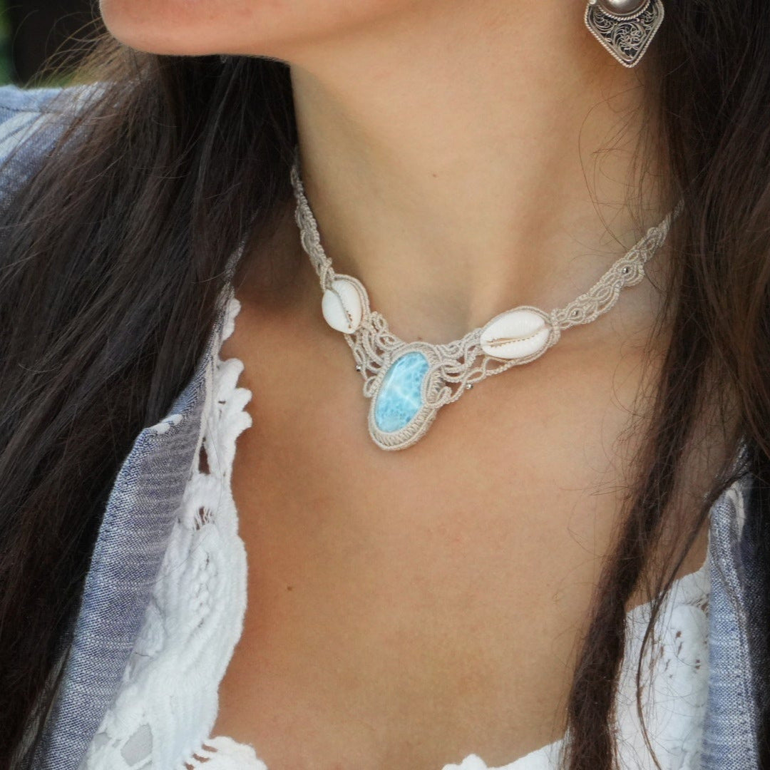 Collier plastron en Larimar Océan bleu ciel, coton couleur lin naturel (beige gris), cauris blancs et perles en acier inoxydable argenté. Système de réglable en tube tissé à l'arrière du bijou. Tissage symétrique en micro macramé avec effet nœuds celtiques entrelacés en relief. Style bohème, hippie, plage, Sirène, tribal et tropical. Convient pour un mariage type bohème ou plage. Bijou porté avec une tenue marine bohème bleu et blanc avec dentelle apparente. Modèle à la peau bronzée et aux cheveux bruns.