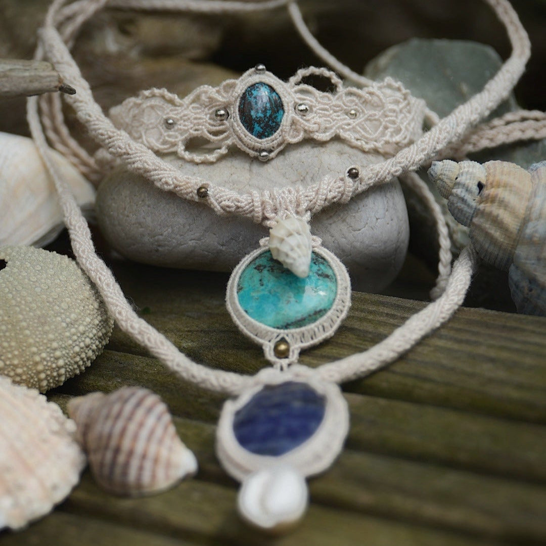 Regroupement de Bijoux en micro macramé ambiance plage et bohème hippie. Bracelet large asymétrique avec tissage dentelle écru et touches argentées. Collier tubulaire torsadé écru avec chrysocolle bleu turquoise et coquillage beige touche de bronze. Collier tubulaire torsadé avec Sodalite bleu foncé, cauri blanc et tissage celtique entrelacé. Bijoux posés sur du bois clair avec éléments marins : coquillages pointus, oursins, galets beige et bois flotté.