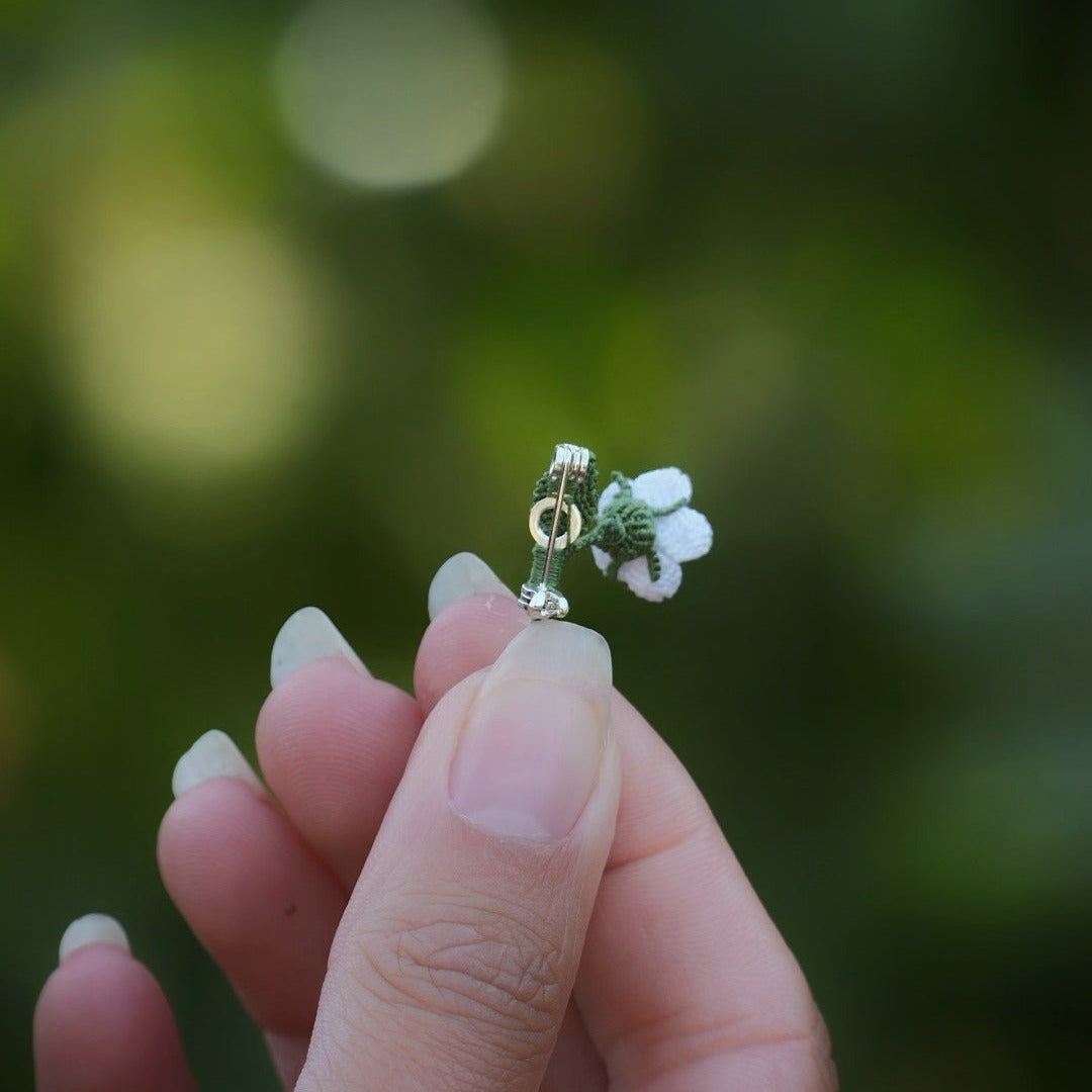 Broche miniature florale blanche en micro macramé avec labradorite bleu-vert au centre, tenue dans une main sur fond de feuilles vertes, bijou bohème et elfique artisanal.