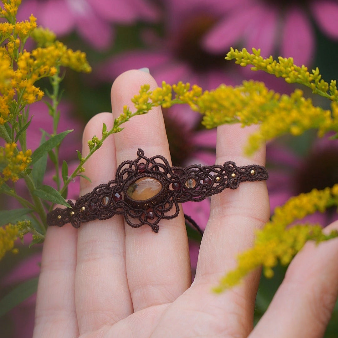 Bracelet réglable en micro macramé. Bijou féminin composé d'une citrine jaune ovale translucide au centre, entourée de 6 perles en grenat rouge à facettes de 3 mm puis de 3 perles dorées de 2 mm. Fil de coton de 0.4 mm tissé en micro macramé avec de motifs en reliefs type nœuds celtiques et motifs arabesques féériques et elfiques. Bijou large ambiance et style bohème hippie, fairycore.
Bijou tenu par une main de femme à la peau claire avec fond vert nature. Vue de face dans un univers floral rose et jaune