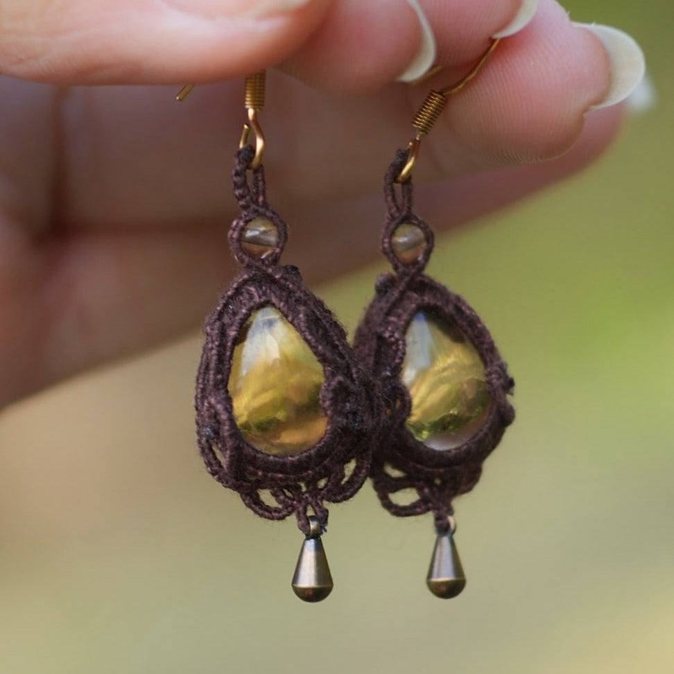 Boucles d'oreilles de style celtique elfique. Bijou féminin composé de : un tissage en relief avec arabesques en micro macramé avec du fil de coton marron foncé chocolat, pierre naturelle de citrine de forme goutte couleur jaune miel accompagné d'une perle ronde en citrine de 3 mm, d'un crochet doré en acier et d'une micro goutte bronze. La paire est présentée de façon verticale tenue entre 2 doigts dans un fond nature vert pâle