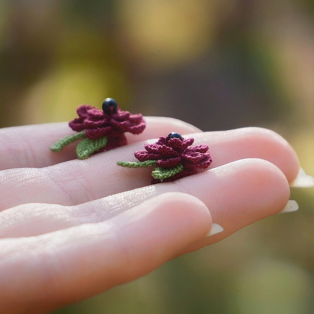 Boucles d’oreilles florales en micro macramé faites main, collection « Pétales éternels », posées sur une main féminine. Fleur 3D tissée en coton rouge bordeaux et vert tendre avec 13 pétales, 2 feuilles et cœur en perle d’onyx noire. Style bohème, elfique et hippie, idéal pour festivals et costumes féériques, photographié au milieu de feuilles et de nature verdoyante.
