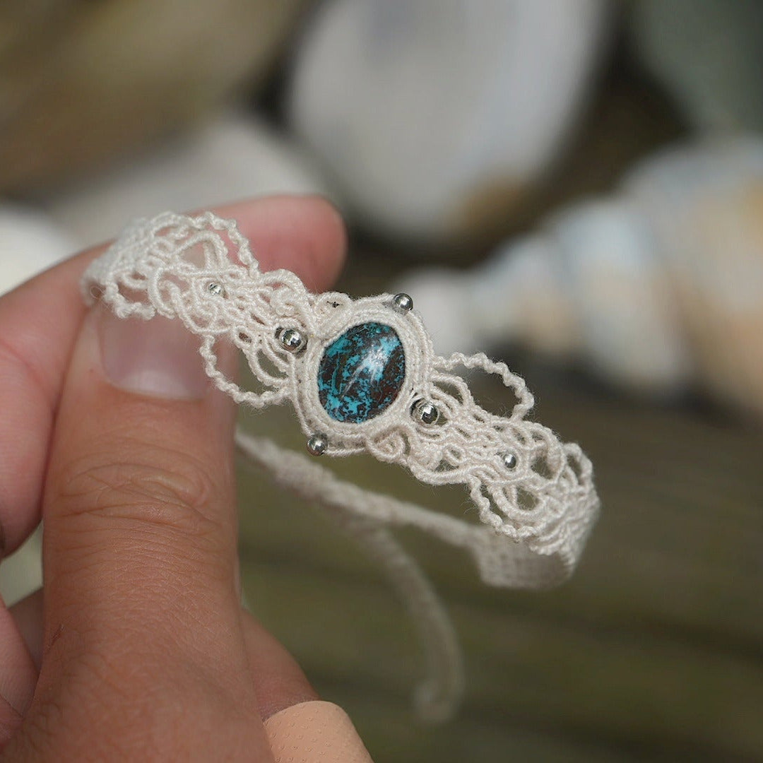 Bracelet large asymétrique. Bijou dentelle ambiance bohème hippie. Bracelet tissé en micro macramé avec fil de coton écru de 0.5 mm avec une pierre ovale de chrysocolle couleur bleu turquoise et marron foncé chocolat. Présence de part et d'autre de façon symétrique des perles en acier inoxydable argentées. Système de réglage à coulisse tubulaire à l'arrière. Design en relief et en torsade entrelacées celtiques. Ambiance plages, festival d'été et mariage. Bijou tenu de face, fond bois clair et coquillages.