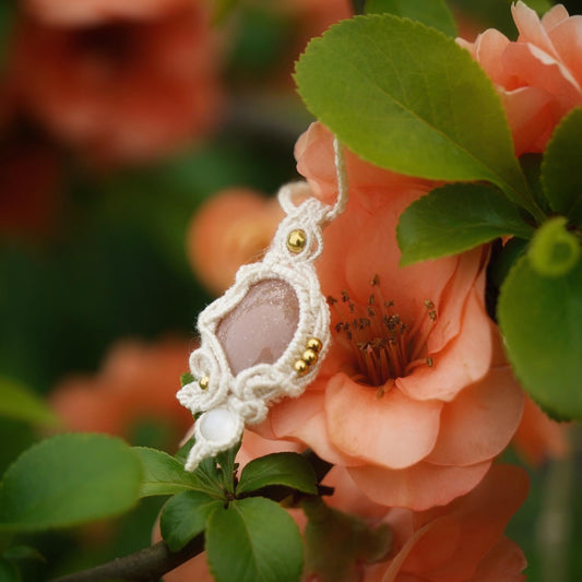 Collier pendentif en micro macramé avec pierre de soleil pêche et nacre, posé sur une branche de cognassier en fleurs saumonées, style bohème et féérique.