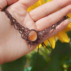 Collier ras-de-cou elfiques en micro macramé avec pierre naturelle. Bijoux tissé en fil de coton de petit diamètre. Mini Grenats rouge rosé facettés autour d'une citrine jaune ambré ovale, fil de coton marron foncé. Motifs tissés en entrelacs celtiques et arabesques elfiques Forme V. Bijoux ambiance bohème hippie aux couleurs chaudes. Bijoux posé à la verticale dans une mains à la peau clair avec fond tournesol jaune et nature. Bijoux vue de face