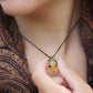 Collier avec pendentif en micro macramé avec motifs floraux. Bijou féminin composé d'un citrine jaune pâle translucide ovale sertie dans une coque marron foncé tissé avec un fil de 0.5 mm sur lequel est fixé une liane verte entrelacée avec mini feuilles et une fleur à 5 pétales jaune orangé avec un cœur doré de 2 mm.

Le bijou est porté autour d'un cou de femme à la peau claire et d'un châle orange et noir. le bijou est tenu entre pouce et index vu de face
