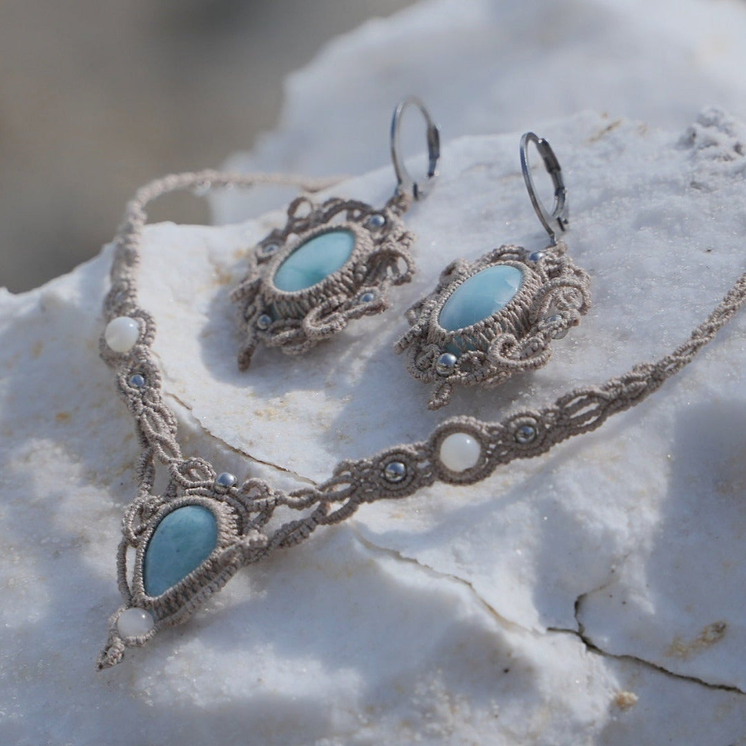Parure Trésors d'Ecume composée d'une paire de boucles d'oreilles pendentes avec collier ras de cou assorti. Larimar bleu ciel ovales pour les boucles et en poire pointe en bas pour le collier. 3 Perles en nacre blanche pour le collier. Perles acier inoxydable argenté pour les deux pour le côté luxe et précieux. Dormeuses en acier inoxydable pour un port assuré. Fil de Coton couleur lin naturel (beige gris) de 0, 5 mm tissé en micro-macramé en relief version celtique. Bijoux posés sur une pierre blanche 