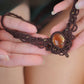 Collier large en micro macramé de couleur marron foncé, jaune ambré et bronze. Motif tissé en micro macramé avec ambiance entrelacs celtiques et arabesques elfiques. Présence d'une pierre centrale ovale de grande taille jaune translucide, gouttes bronze, forme V avec mini perles ronde bronze parsemées. Photo ambiance bohème hippie avec une touche d'élégance. Bijou posé à plat dans 2 mains en coupe à peau claire.  jupe vert forêt en velours côtelé à l'arrière