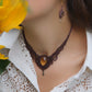 Collier large en micro macramé de couleur marron foncé, jaune ambré et bronze. Motif tissé en micro macramé avec ambiance entrelacs celtiques et arabesques elfiques. Présence d'une pierre centrale ovale de grande taille jaune translucide, gouttes bronze, forme V avec mini perles ronde bronze parsemées. Photo ambiance bohème hippie avec une touche d'élégance. Bijou porté sur une peau claire avec tournesol jaune en premier plan et chemise blanche en dentelle.