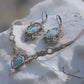 Parure Trésors d'Ecume composée d'une paire de boucles d'oreilles pendentes avec collier ras de cou assorti. Larimar bleu ciel ovales pour les boucles et en poire pointe en bas pour le collier. 3 Perles en nacre blanche pour le collier. Perles acier inoxydable argenté pour les deux pour le côté luxe et précieux. Dormeuses en acier inoxydable pour un port assuré. Fil de Coton couleur lin naturel (beige gris) de 0, 5 mm tissé en micro-macramé en relief version celtique. Bijoux posés sur une pierre blanche 