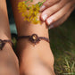 Bijoux de pieds, paire assortie de bracelets de chevilles à ambiance bohème celtique elfique. Fil marron foncé fin tissé en micro macramé avec entrelacs et arabesques, pierre centrale ovale petite taille jaune miel translucide. Bijoux hippie, bohème avec inspiration nature. Chevillères portées sur des chevilles à la peau pâle, bijoux de face avec jeu d'ombre et lumière et fleurs jaune en haut à droite.
