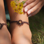 Bijoux de pieds, paire assortie de bracelets de chevilles à ambiance bohème celtique elfique. Fil marron foncé fin tissé en micro macramé avec entrelacs et arabesques, pierre centrale ovale petite taille jaune miel translucide. Bijoux hippie, bohème avec inspiration nature. Chevillères portées sur des chevilles à la peau pâle, bijoux de face avec jeu d'ombre et lumière et fleurs jaune en haut à droite.