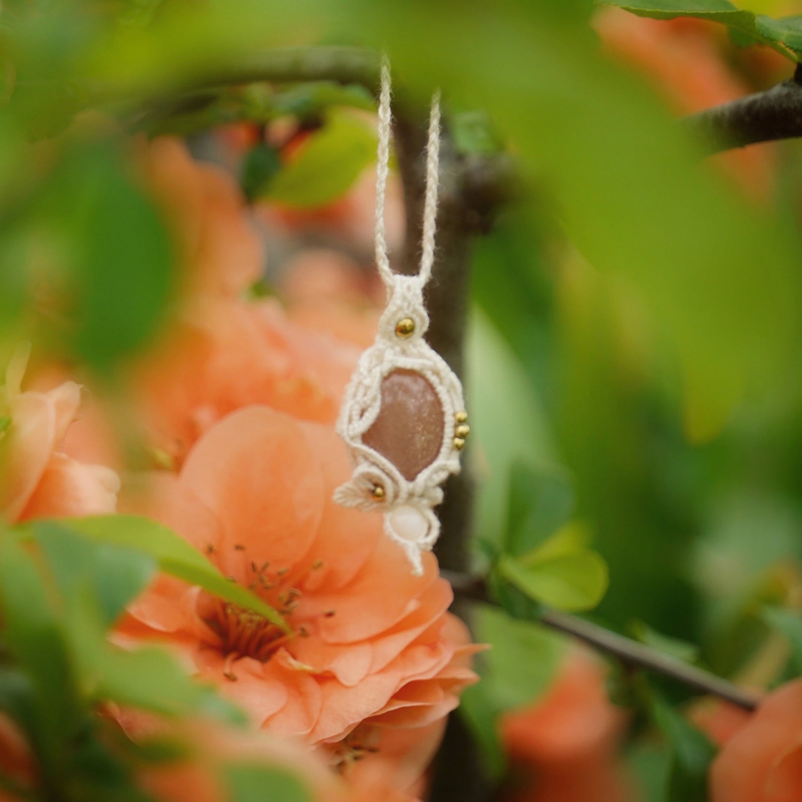 Collier pendentif en micro macramé avec pierre de soleil pêche et nacre, posé sur une branche de cognassier en fleurs saumonées, style bohème et féérique.