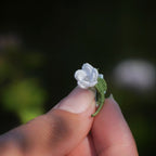 Broche miniature florale blanche en micro macramé avec labradorite bleu-vert au centre, tenue dans une main sur fond de feuilles vertes, bijou bohème et elfique artisanal.