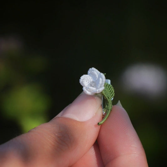 Broche miniature florale blanche en micro macramé avec labradorite bleu-vert au centre, tenue dans une main sur fond de feuilles vertes, bijou bohème et elfique artisanal.