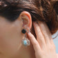Boucles d'oreilles Trésors d'Ecume composée d'une Larimar bleu ciel ovales, mini Perles rondes en acier inoxydable argenté pour le côté luxe et précieux. Dormeuses en acier inoxydable pour un port assuré. Fil de Coton couleur lin naturel (beige gris) de 0, 5 mm tissé en micro-macramé en relief version celtique. Collage 3 photos : Bijoux posés et porté sur une femme bronzée et brune, et descriptif de composition visuel avec flèche 