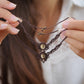 Bijoux de pieds, paire assortie de bracelets de chevilles à ambiance bohème celtique elfique. Fil marron foncé fin tissé en micro macramé avec entrelacs et arabesques, pierre centrale ovale petite taille jaune miel translucide. Bijoux hippie, bohème avec inspiration nature. Chevillères tenues entre 2 mains à la peau pâle l'une au dessus de l'autre en verticale avec en fond une femme aux cheveux bruns et avec une chemise blanche