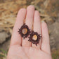 Paire de boucles d'oreilles et d'un collier ras-de-cou elfiques en micro macramé avec pierre naturelle. Bijoux tissé en fil de coton de petit diamètre. Mini Grenats rouge rosé facettés autour d'une citrine jaune ambré, fil de coton marron foncé. Motifs tissés en entrelacs celtiques et arabesques elfiques. Crochet doré. Bijoux ambiance bohème hippie aux couleurs chaudes. Bijoux posés, vue d'en haut dans une main à la peau claire.