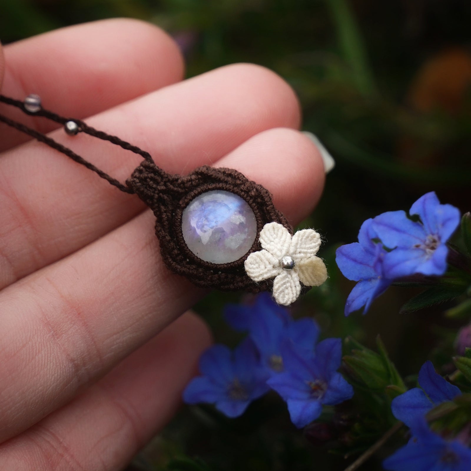Collier artisanal en labradorite blanche tenu dans une main près de fleurs bleu cobalt, bijou elfique poétique inspiré de la pluie, micro macramé coton et perles fines. Coton et Astrance