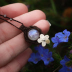 Collier artisanal en labradorite blanche tenu dans une main près de fleurs bleu cobalt, bijou elfique poétique inspiré de la pluie, micro macramé coton et perles fines. Coton et Astrance