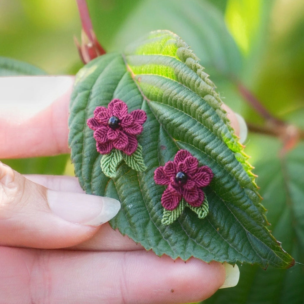 Boucles d’oreilles florales en micro macramé faites main, collection « Pétales éternels », posées sur une main féminine. Fleur 3D tissée en coton rouge bordeaux et vert tendre avec 13 pétales, 2 feuilles et cœur en perle d’onyx noire. Style bohème, elfique et hippie, idéal pour festivals et costumes féériques, photographié au milieu de feuilles et de nature verdoyante.