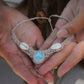 Collier plastron en Larimar Océan bleu ciel, coton couleur lin naturel (beige gris), cauris blancs et perles en acier inoxydable argenté. Système de réglable en tube tissé à l'arrière du bijou. Tissage symétrique en micro macramé avec effet nœuds celtiques entrelacés en relief. Style bohème, hippie, plage, Sirène, tribal et tropical. Convient pour un mariage type bohème ou plage. Bijou tenu vue 3/4, tenue romantique et bohème blanche avec dentelle apparente. Modèle à la peau bronzée et aux cheveux bruns. 