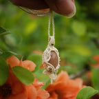 Collier pendentif en micro macramé avec pierre de soleil pêche et nacre, posé sur une branche de cognassier en fleurs saumonées, style bohème et féérique.