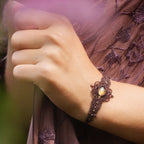 Bracelet large elfique en micro macramé brun, orné d’une citrine jaune pâle et de perles de grenat rouge rosé. Bijou bohème et artisanal réglable, porté au poignet d’une femme à la peau claire.